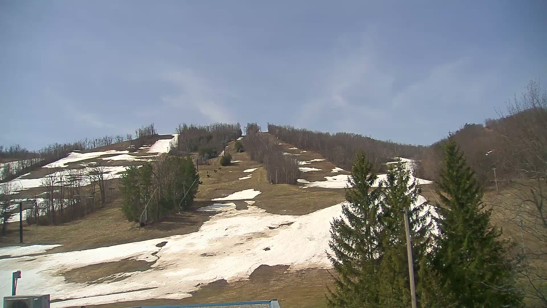 A mostly clear, sunny day reveals a ski slope with patches of melting snow and brown grass, surrounded by leafless trees.