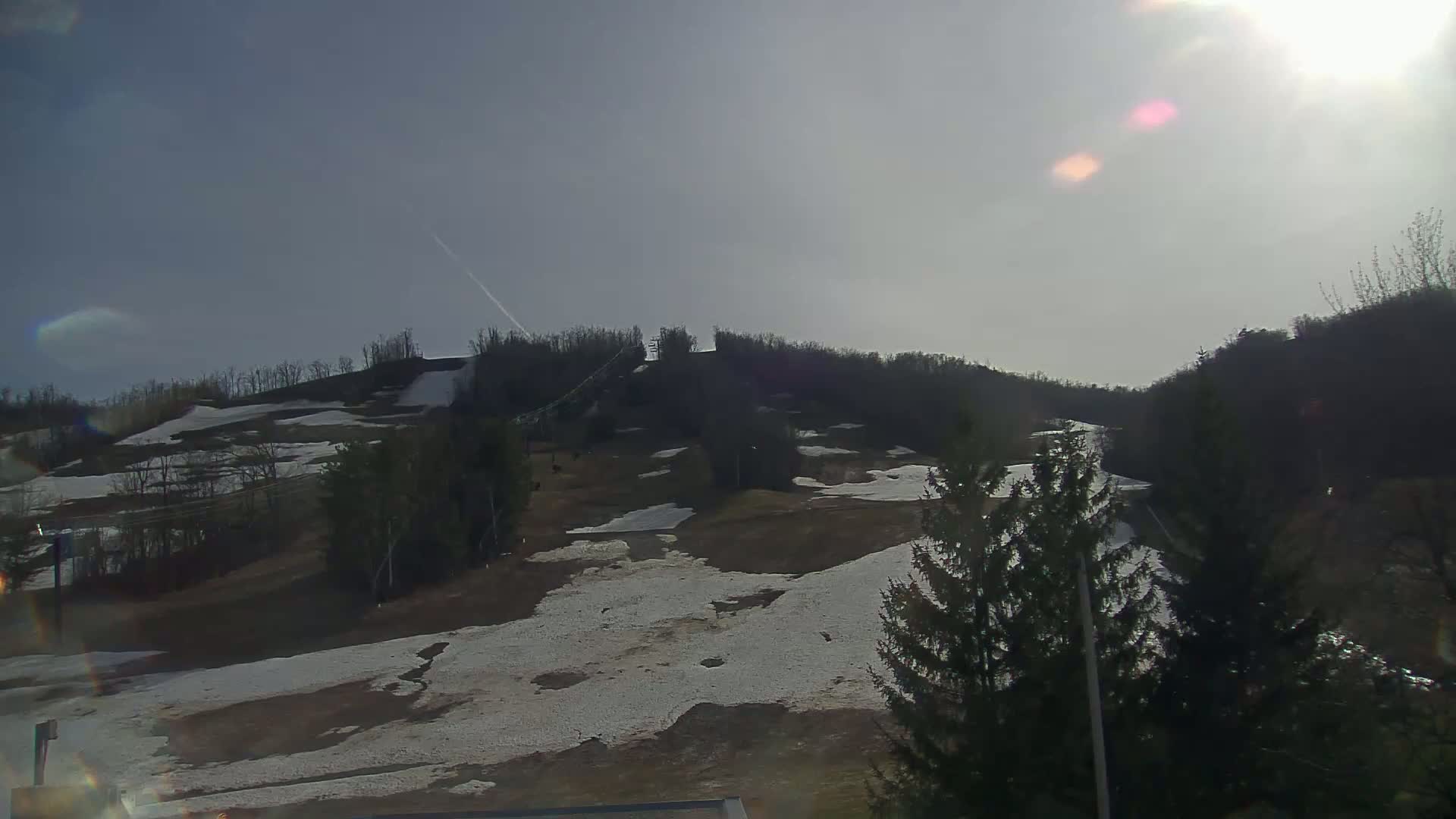 A mostly sunny day reveals a snow-covered hillside with patches of brown earth, trees, and ski lift infrastructure.