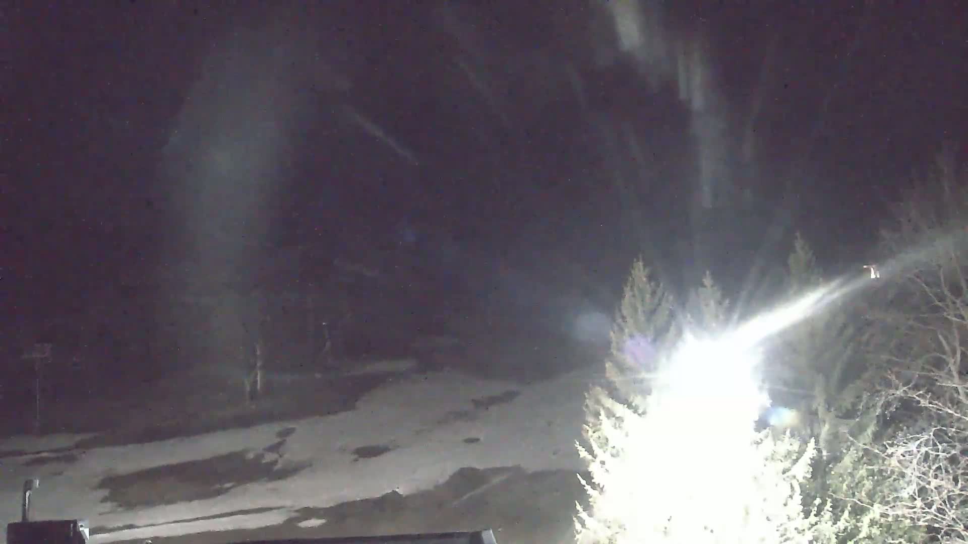 A nighttime scene of snow falling over a snow-covered landscape with trees and bright lights illuminating the area.