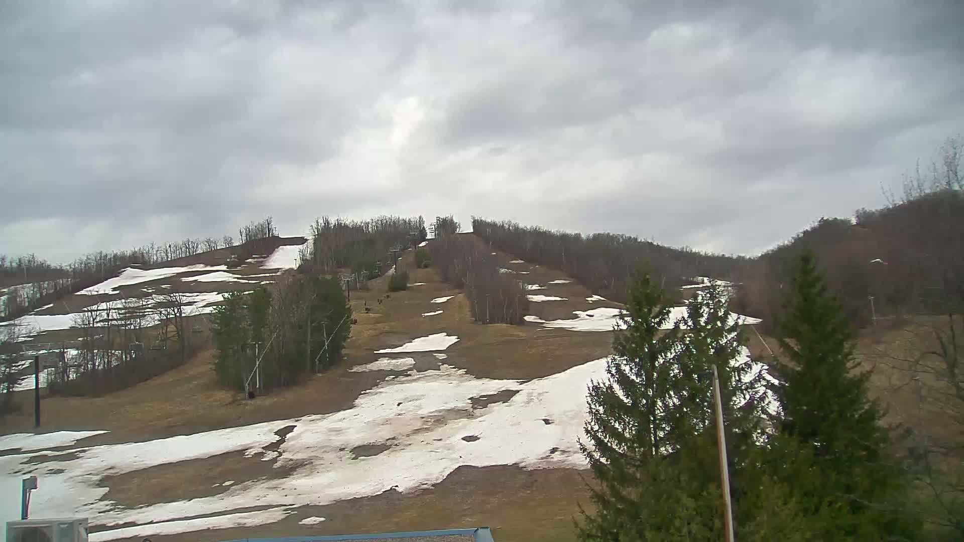 A mostly cloudy day reveals a ski hill with patches of melting snow, surrounded by bare trees.