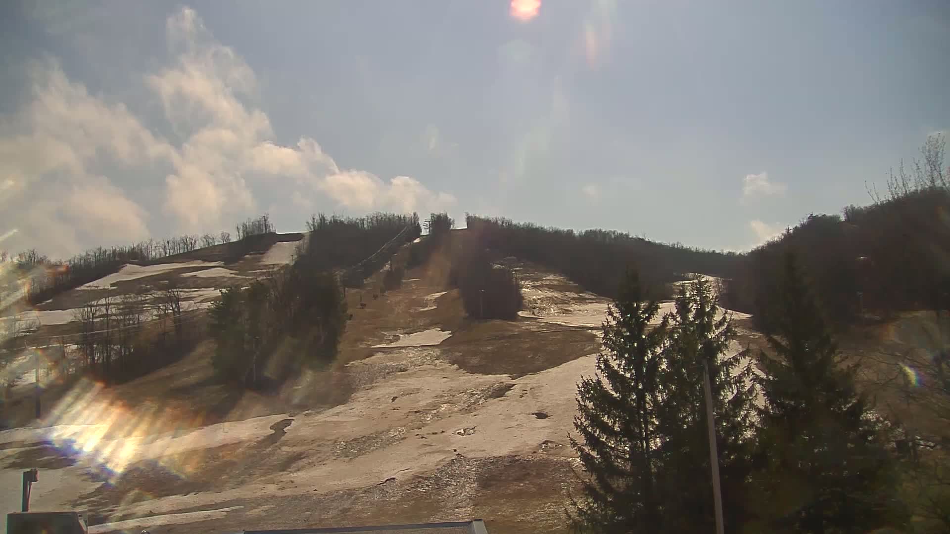 A mostly sunny day shows a snow-covered ski slope with patches of bare ground, surrounded by leafless trees.