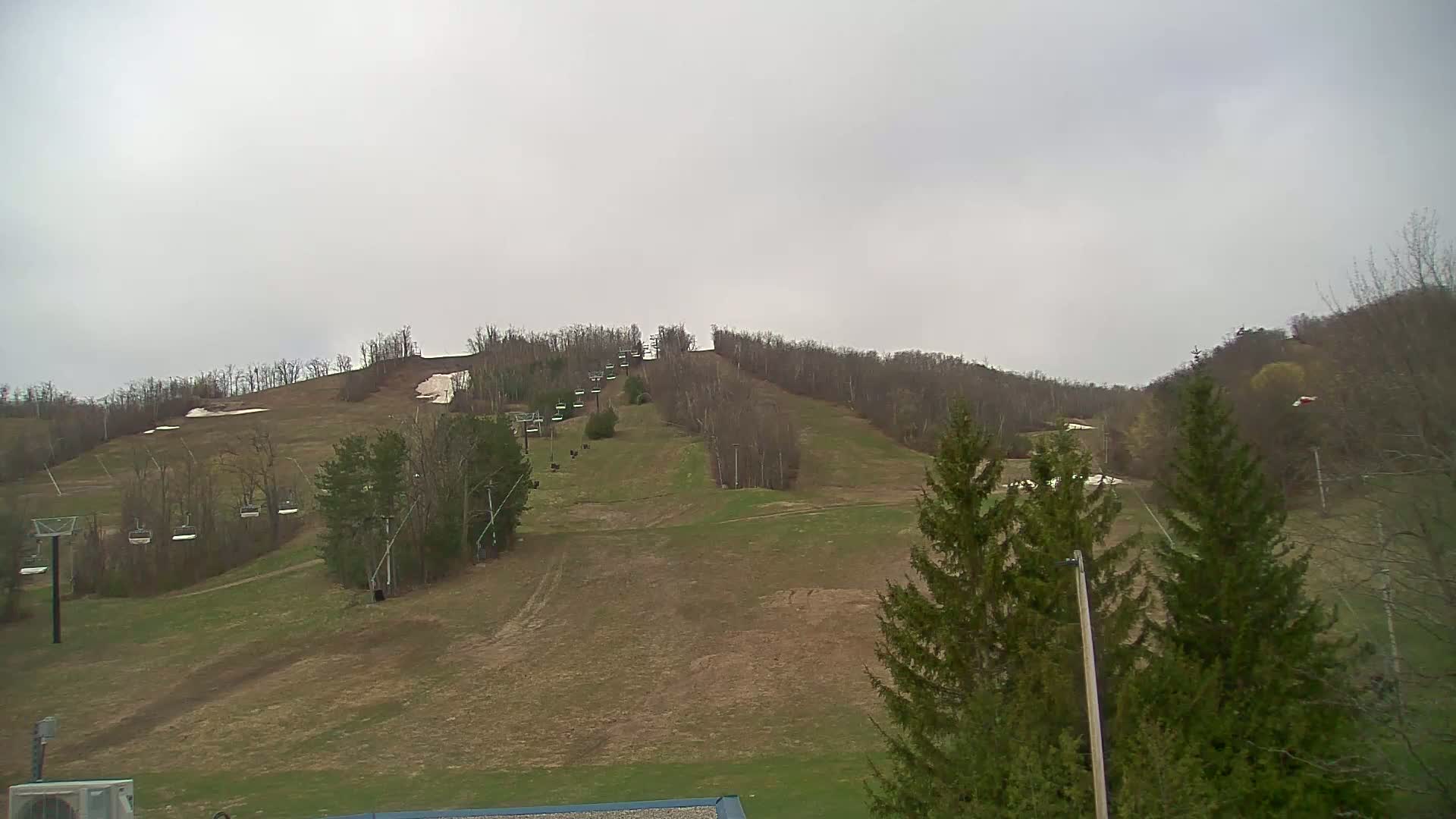 A ski slope with a chairlift, mostly brown grass, and some remaining snow patches under an overcast sky.
