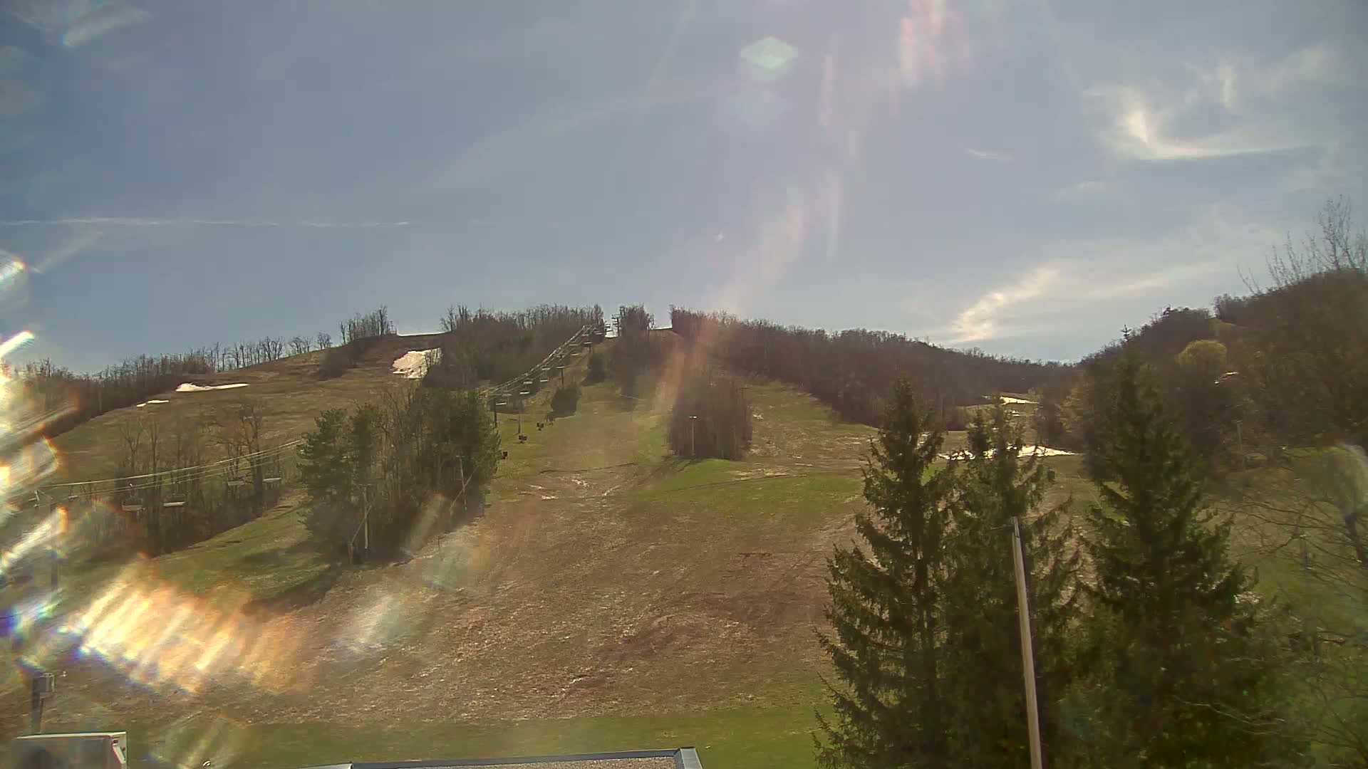 A mostly sunny day shows a grassy hillside with a ski lift and patches of remaining snow.