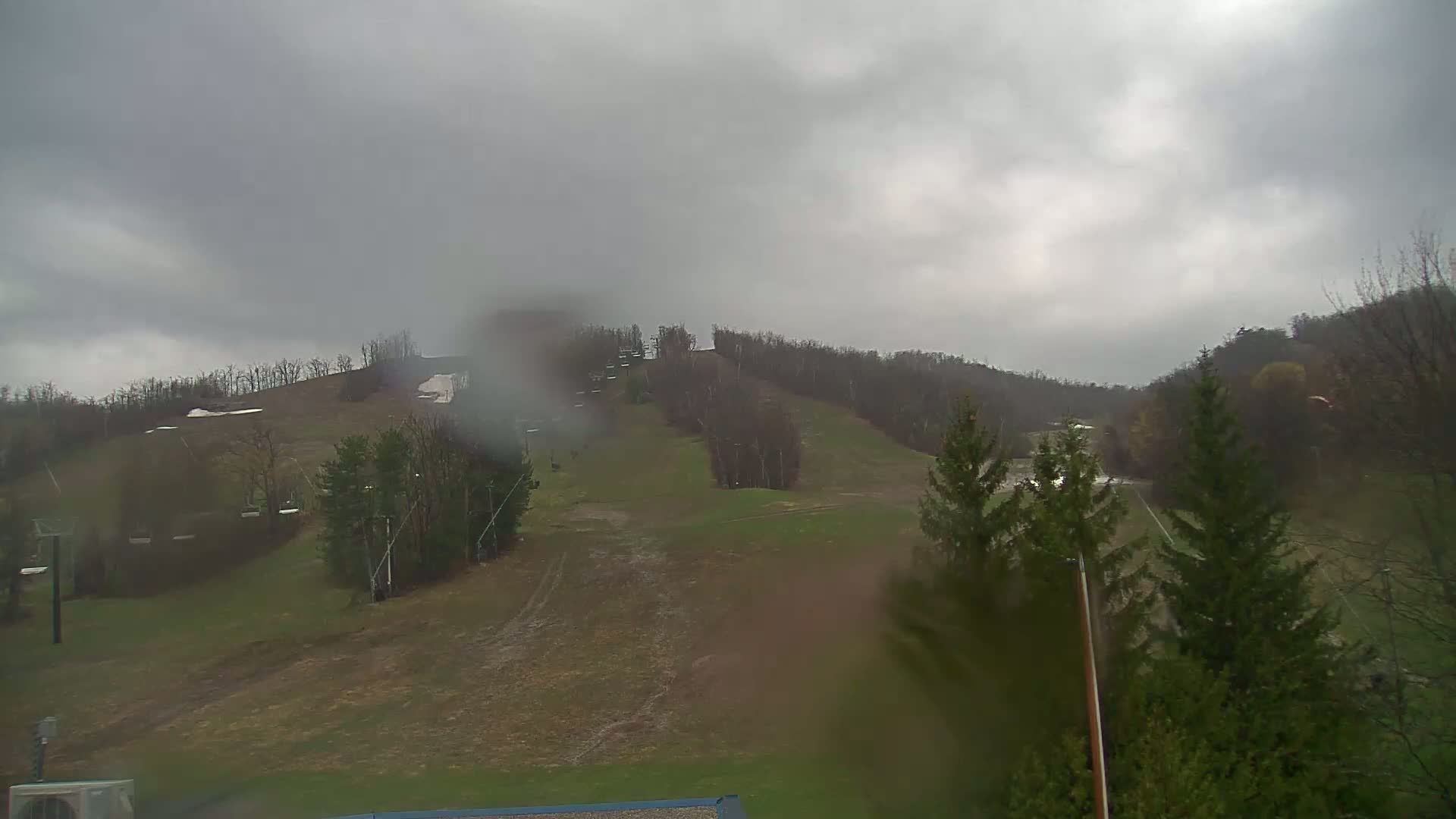 A view of a ski slope with some remaining snow patches under an overcast, rainy sky.