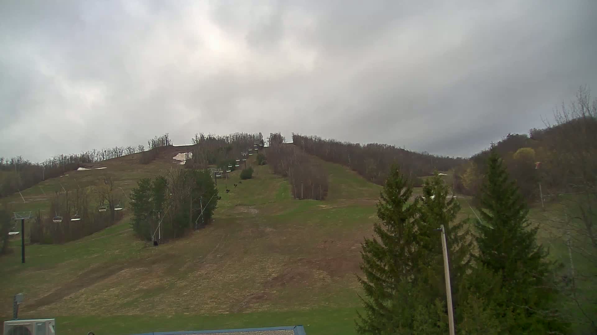 A mostly cloudy day reveals a ski hill with chairlifts and patchy snow, surrounded by early spring foliage.