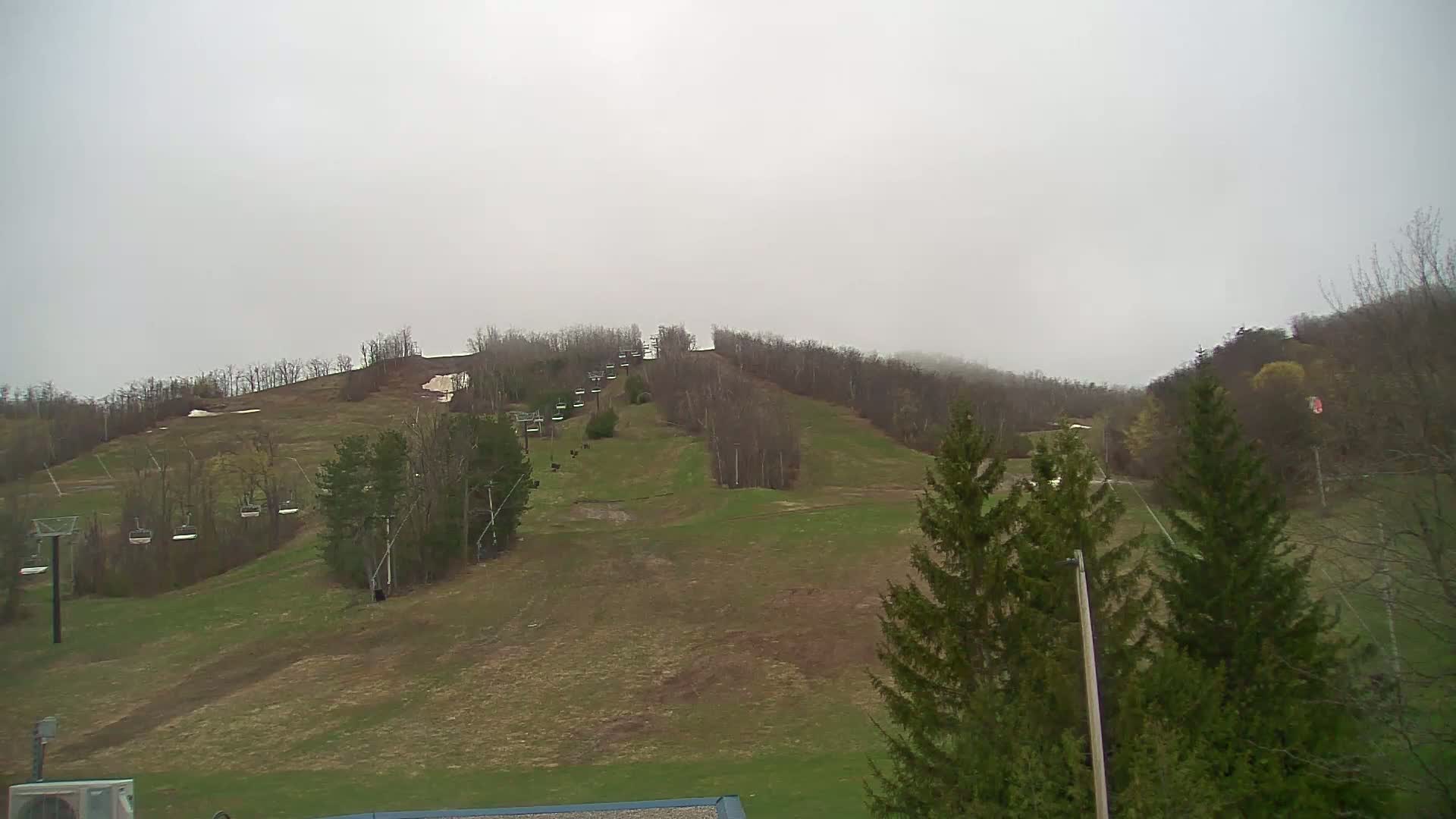 A ski slope with chairlifts is visible under an overcast sky, showing patches of remaining snow and early spring vegetation.