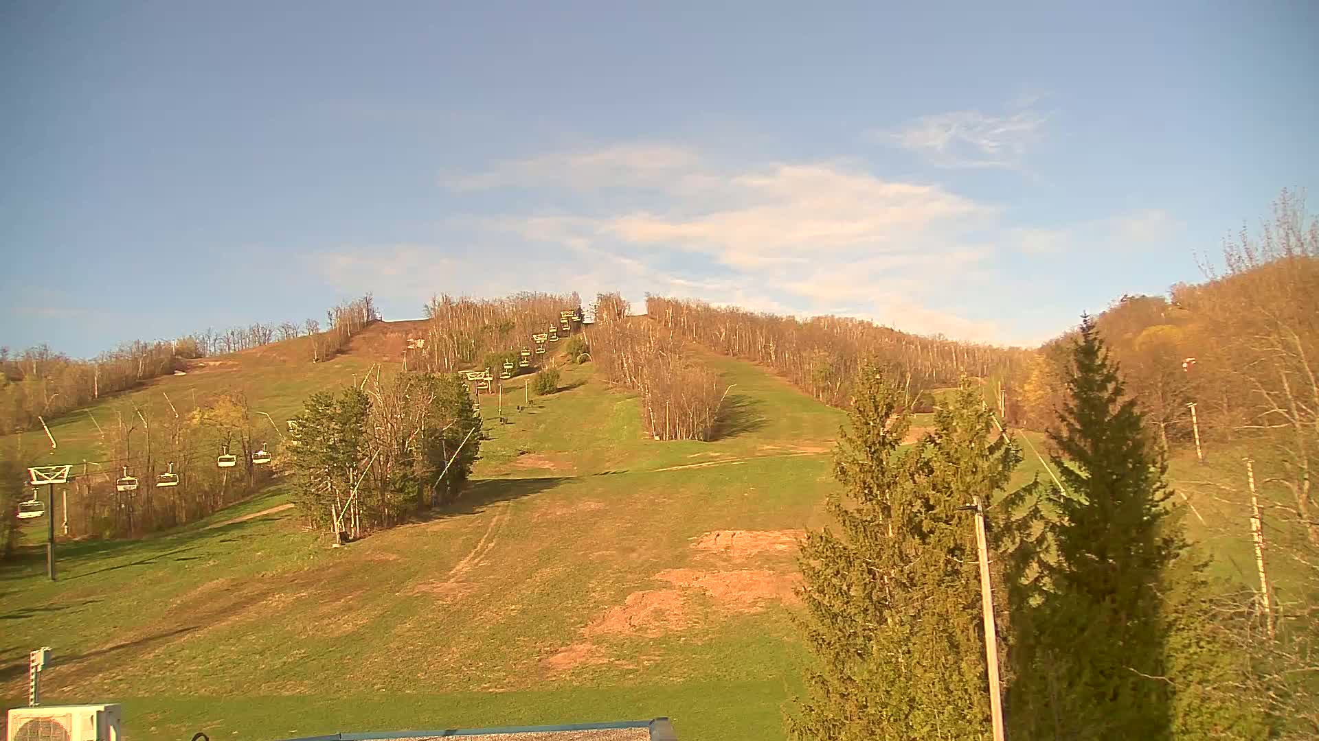 A grassy ski hill with a chairlift and mostly bare trees under a mostly clear, sunny sky.