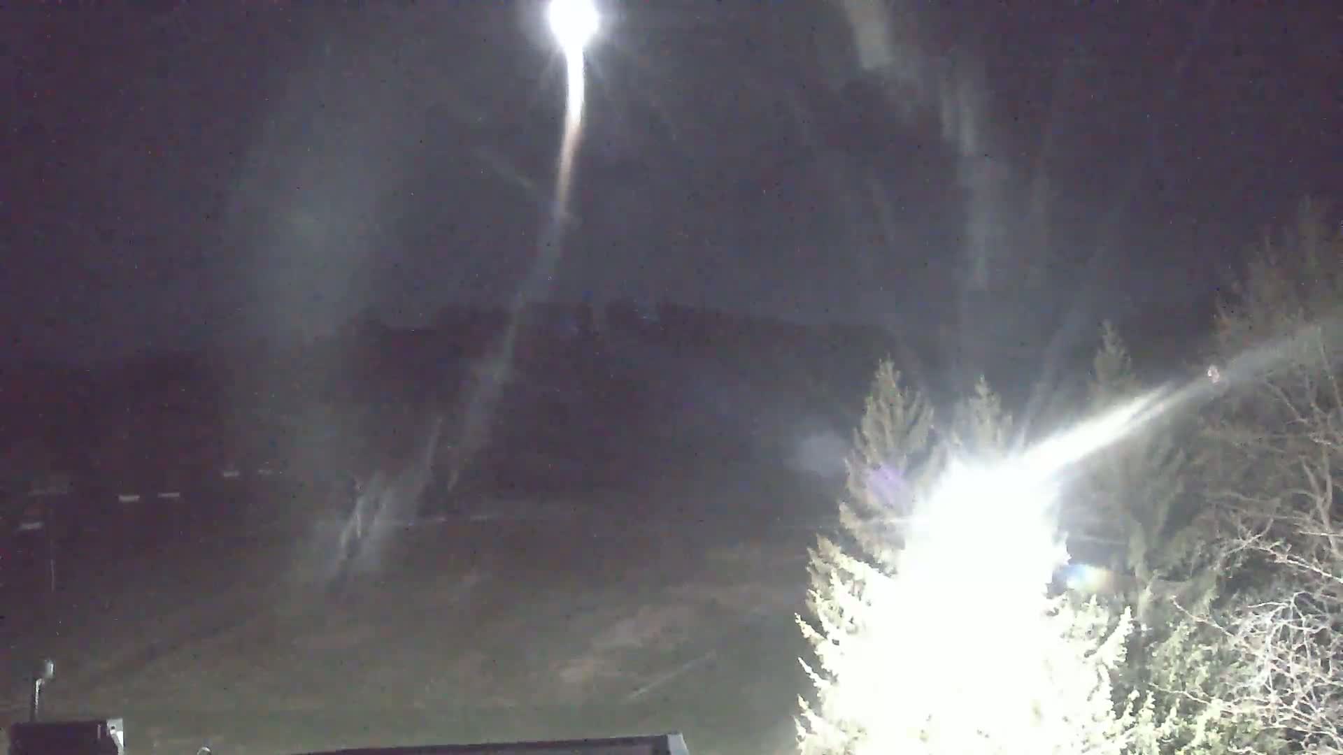Bright light streaks across a nighttime sky above a dark, snow-covered landscape with silhouetted trees.