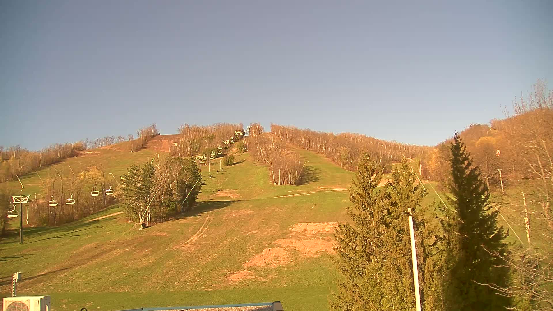 A grassy ski slope with chairlifts and sparse, early spring trees under a clear, light-blue sky.