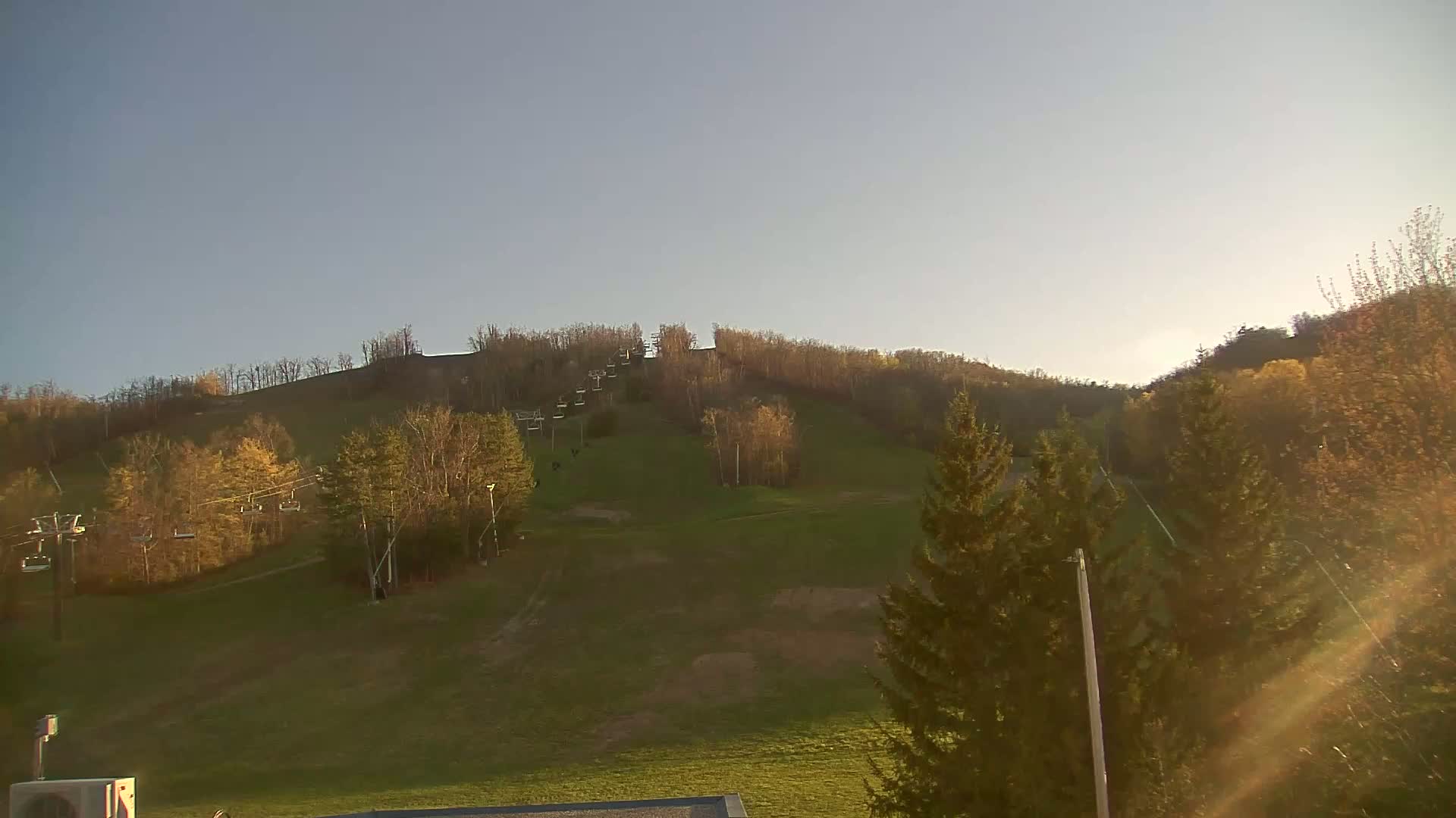 A grassy hill with a chairlift and sparsely leaved trees under a clear, light blue sky.