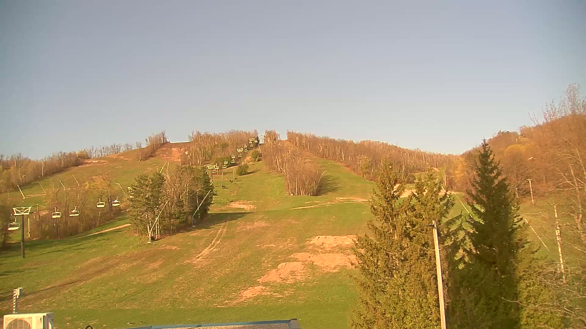 A grassy ski hill with a chairlift and sparse, early spring deciduous trees under a clear, light blue sky.