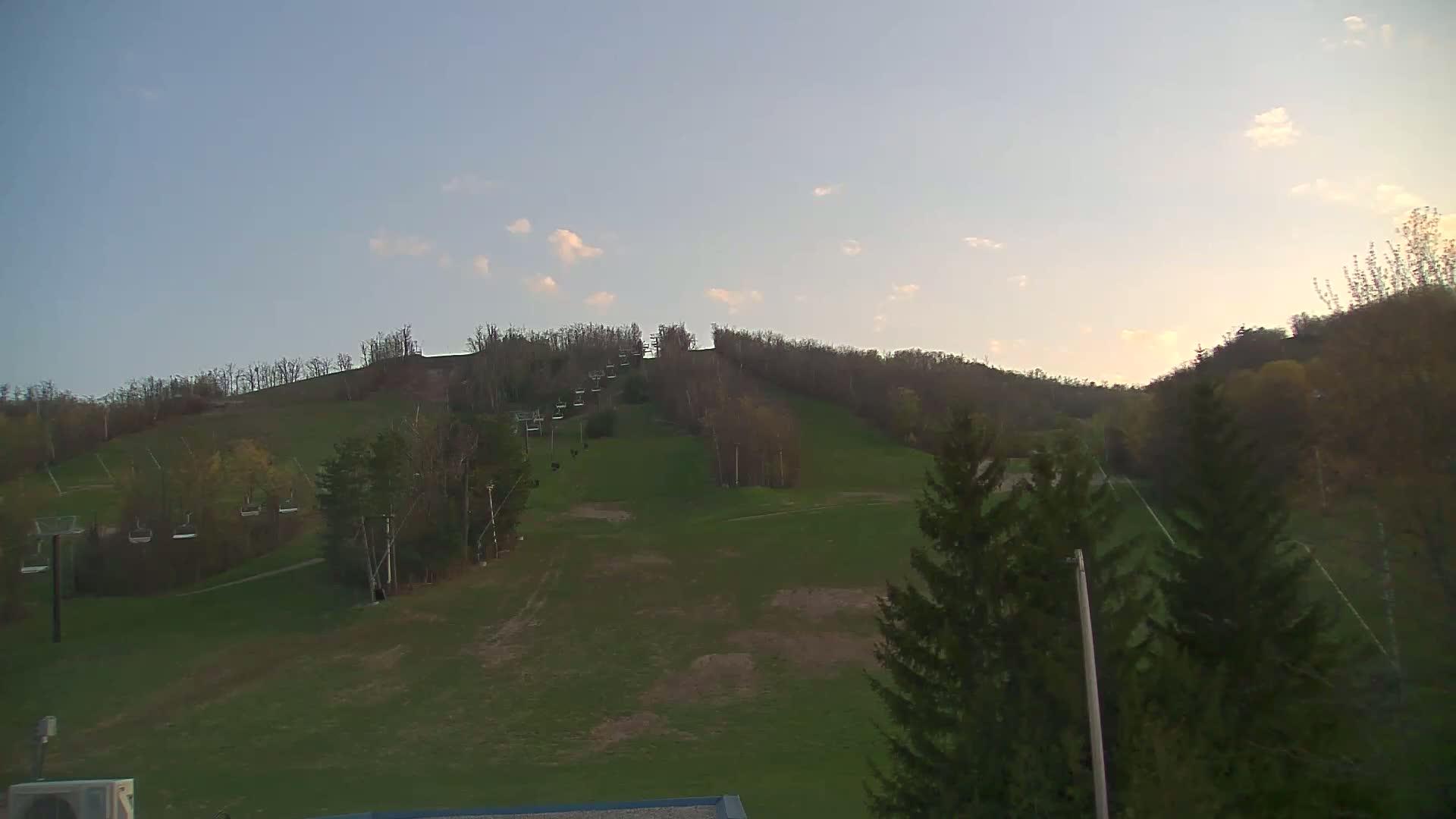 A grassy hillside with a chairlift and sparse trees under a mostly clear, light blue sky.