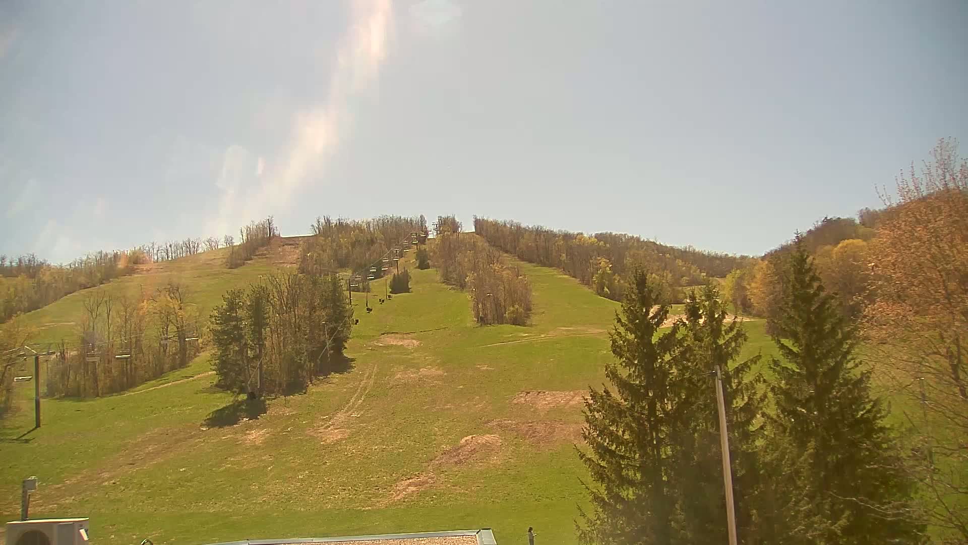 A grassy ski slope with a chairlift and sparse trees under a mostly sunny sky.