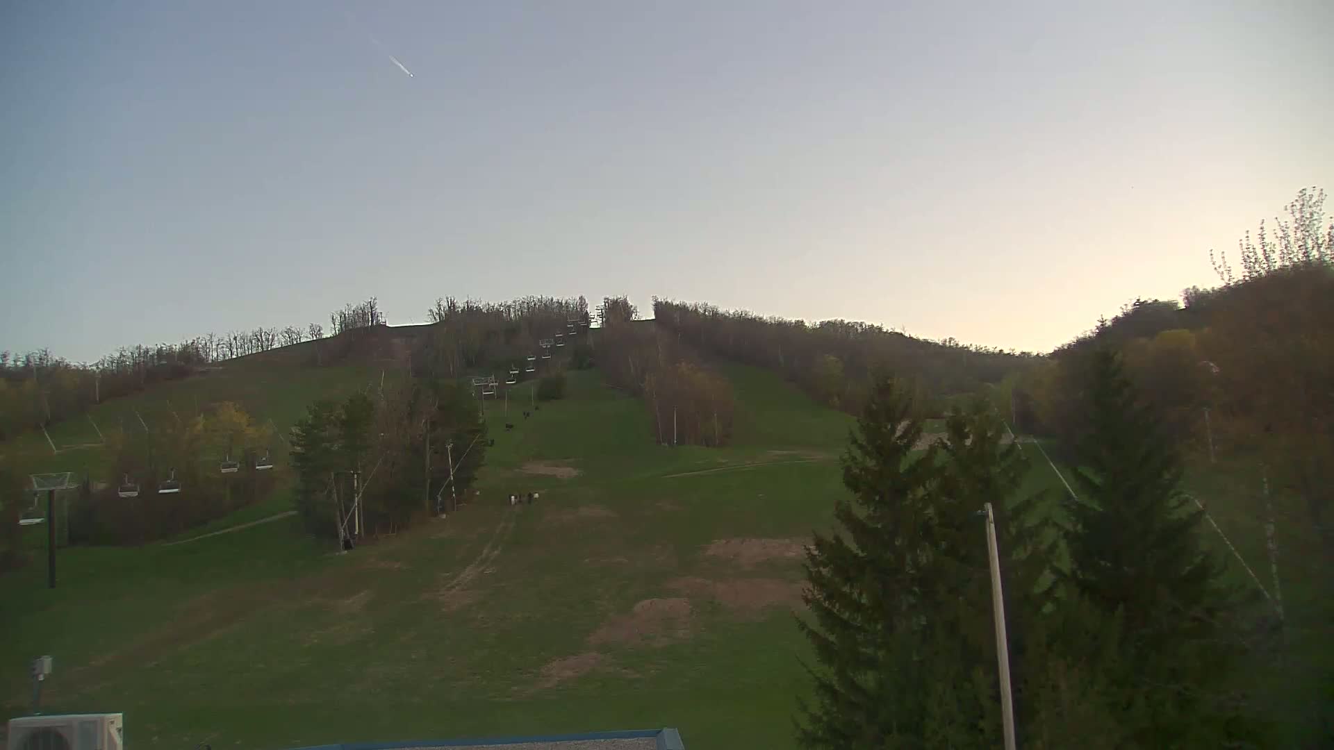 A grassy hillside with a ski lift and several trees under a clear, light-blue sky.