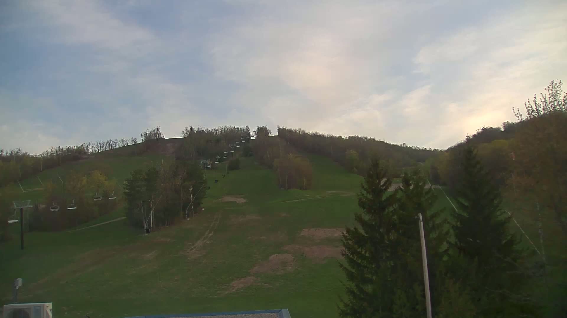 A grassy hill with a chairlift and deciduous trees under a mostly cloudy sky.