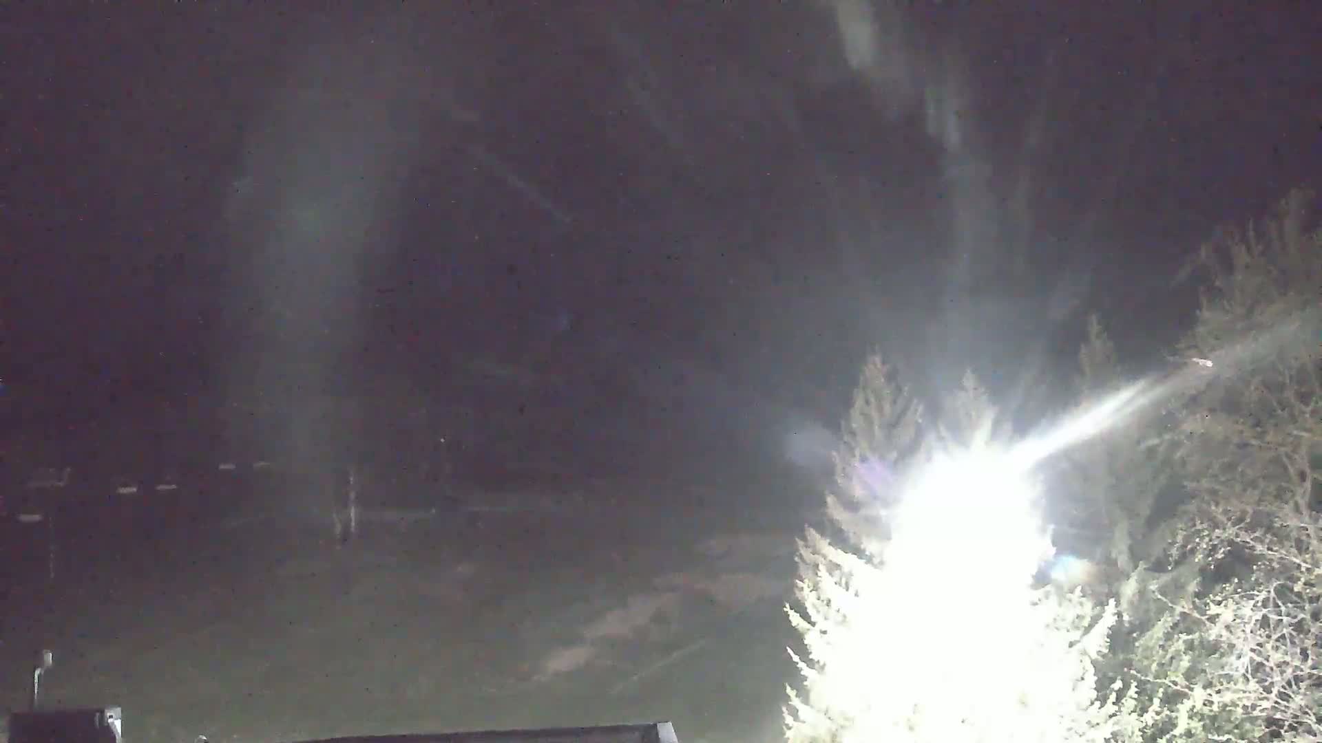 A bright light shines through bare trees at night, illuminating a dark, snowy landscape.