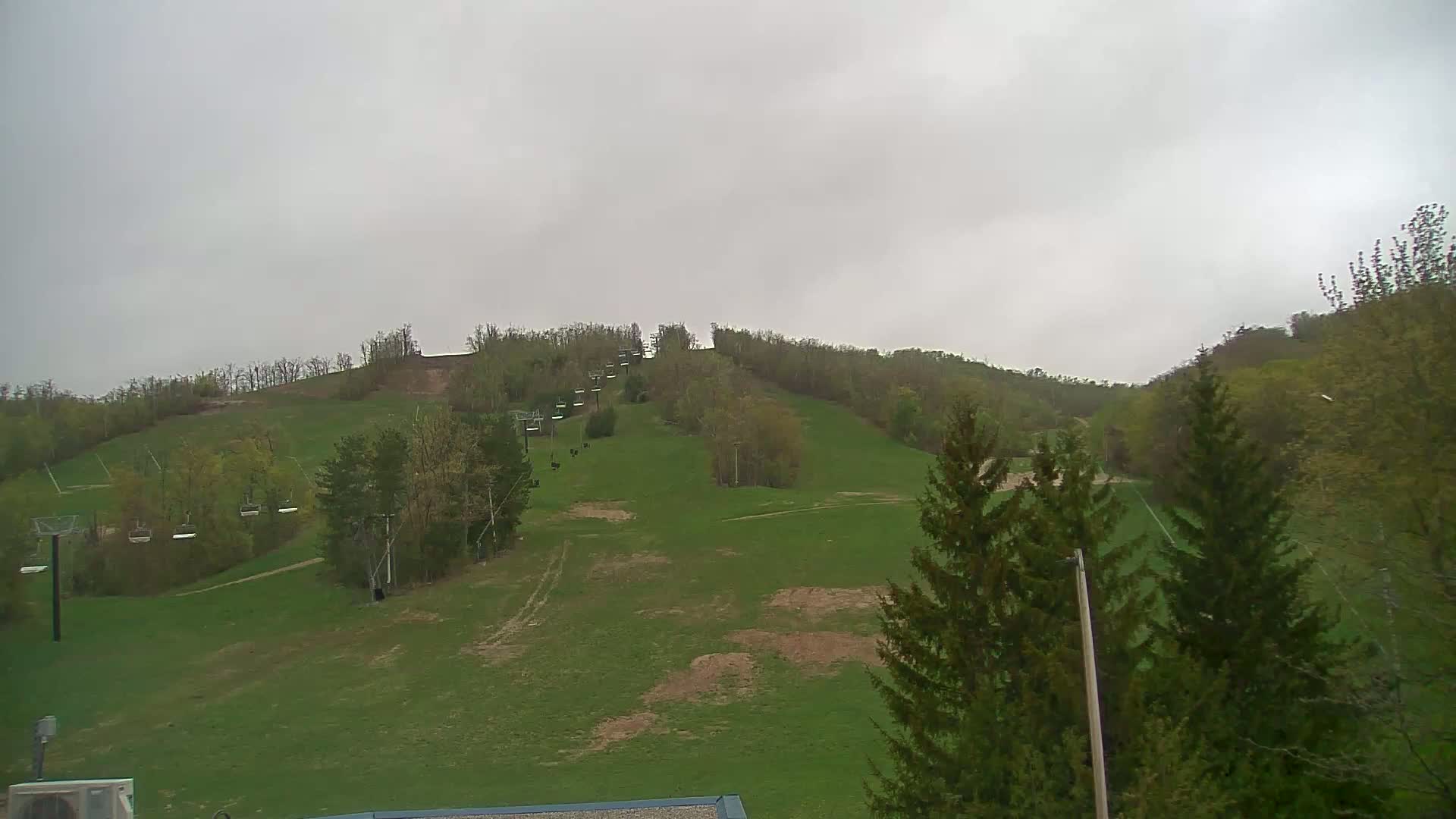 A grassy hillside with a chairlift and surrounding trees is visible under an overcast sky.