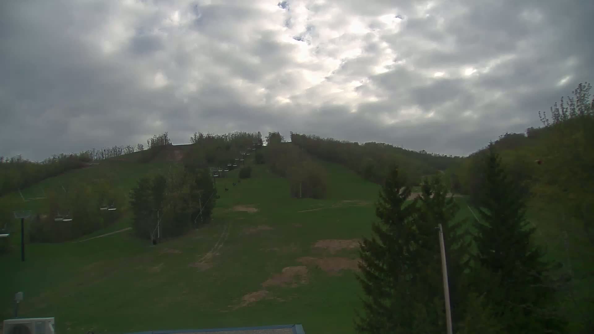 A grassy hill with several chairlifts and trees under a cloudy sky.