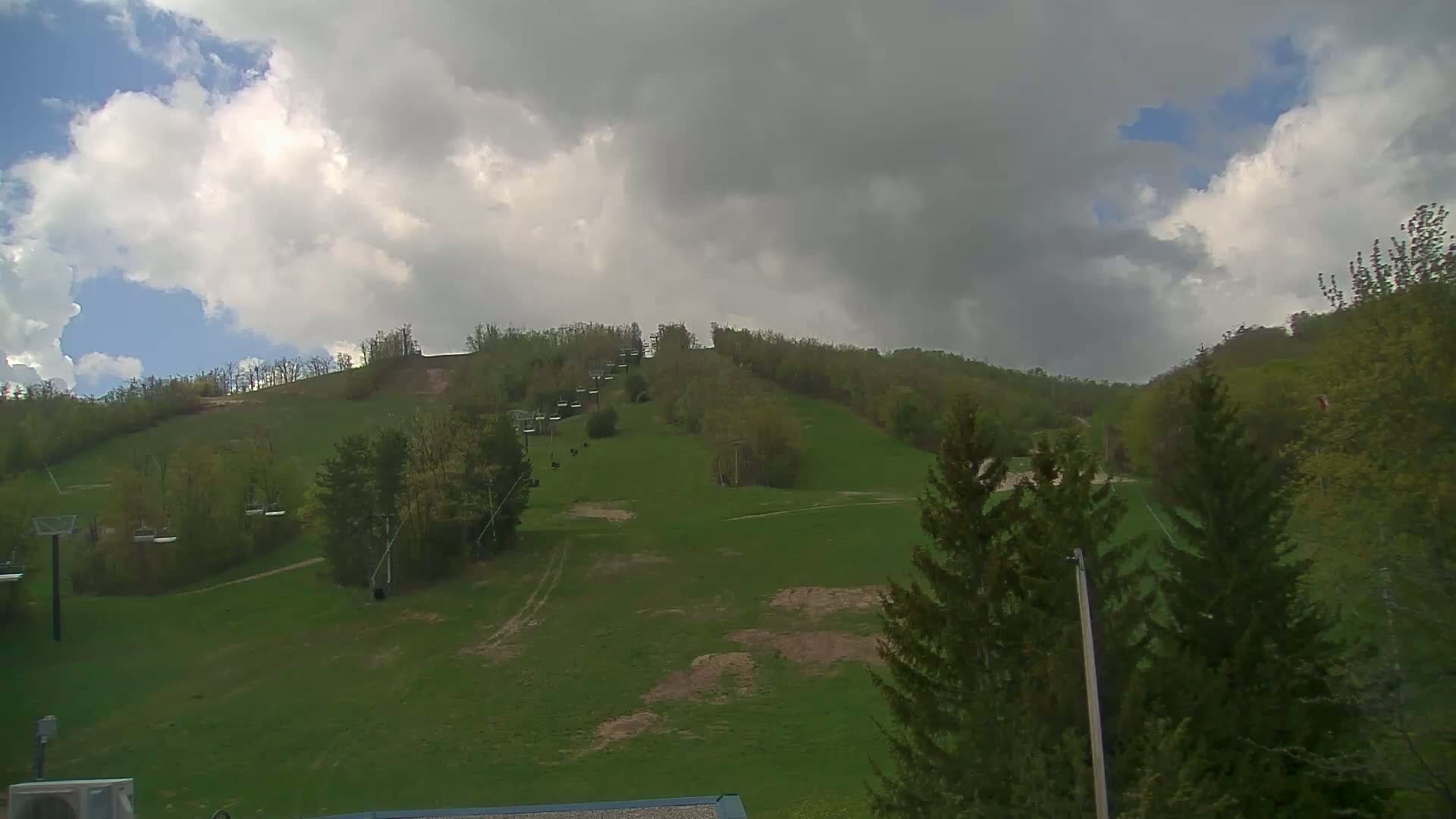 A grassy ski hill with chairlifts and some trees under a partly cloudy sky.