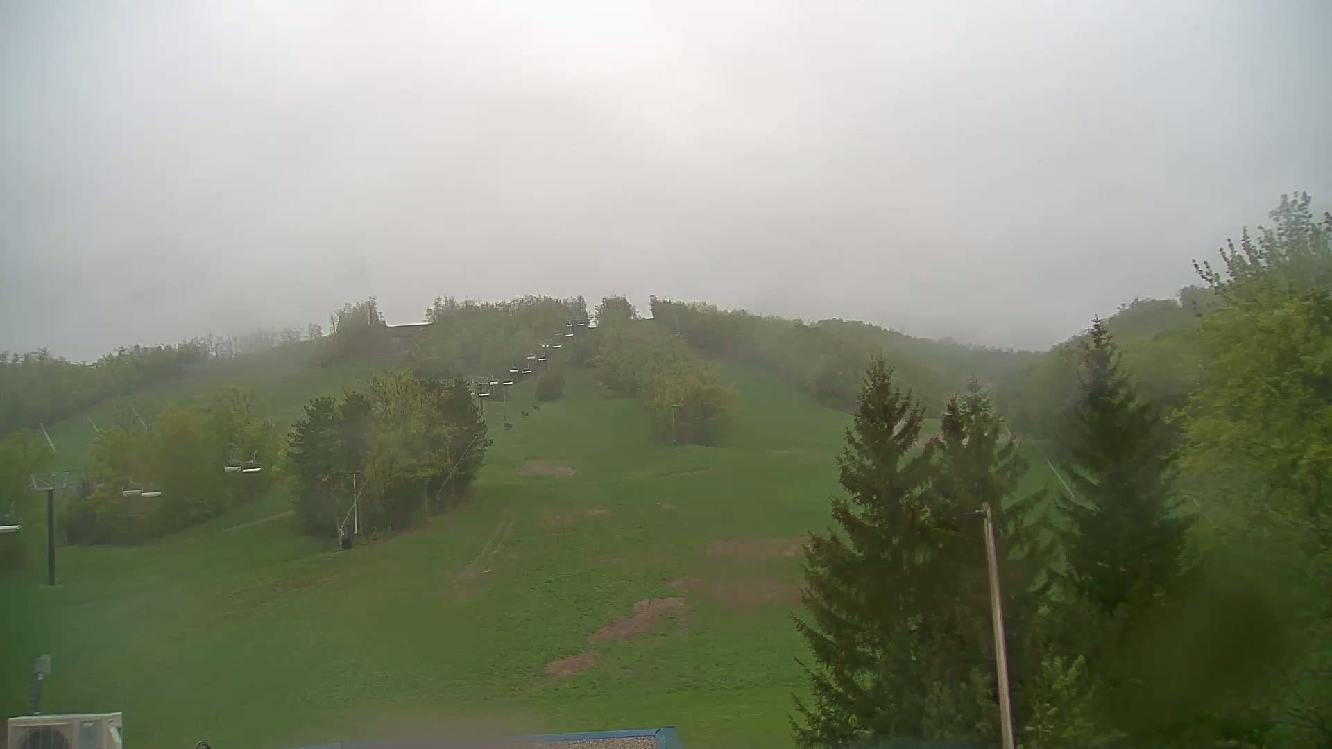 A grassy hillside with a chairlift and trees is visible in foggy, rainy weather.