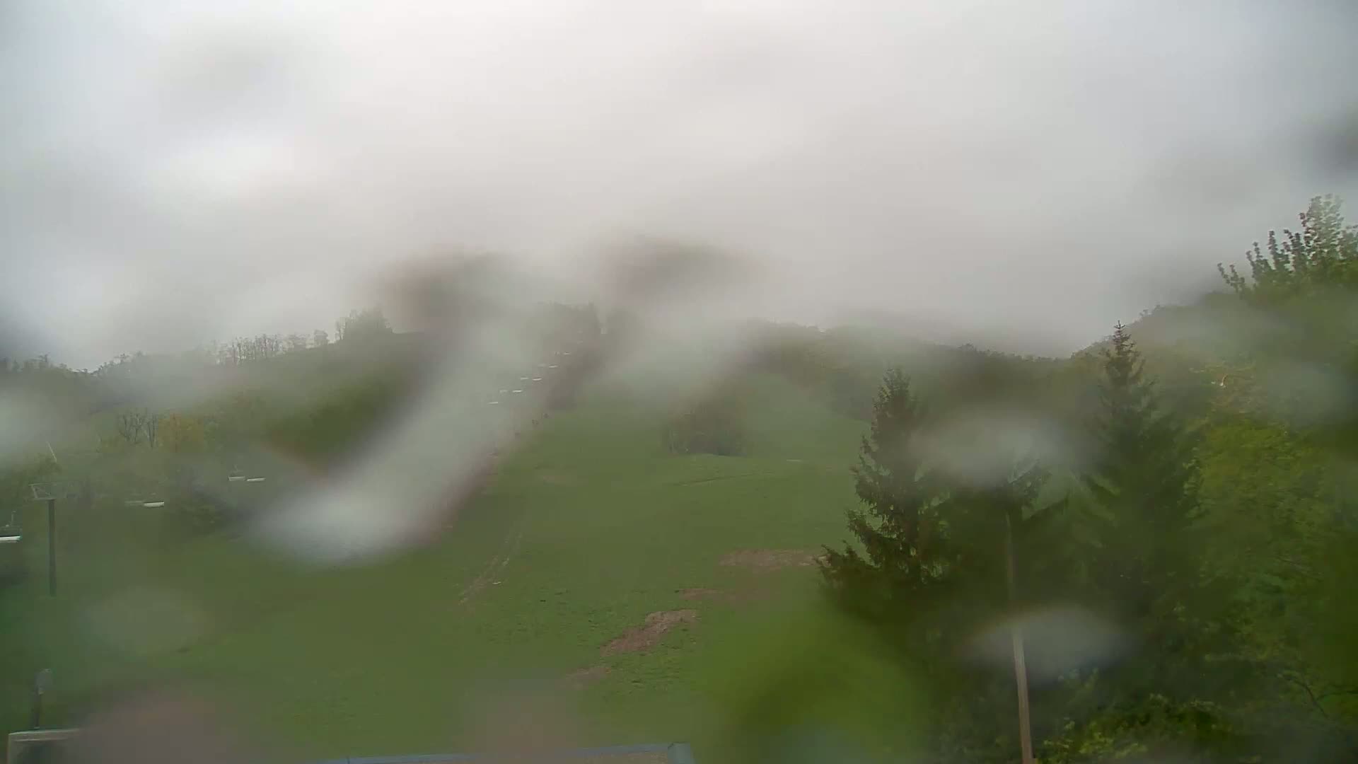 A rain-covered view of a grassy hillside with trees and what appear to be ski lifts in the distance under an overcast sky.
