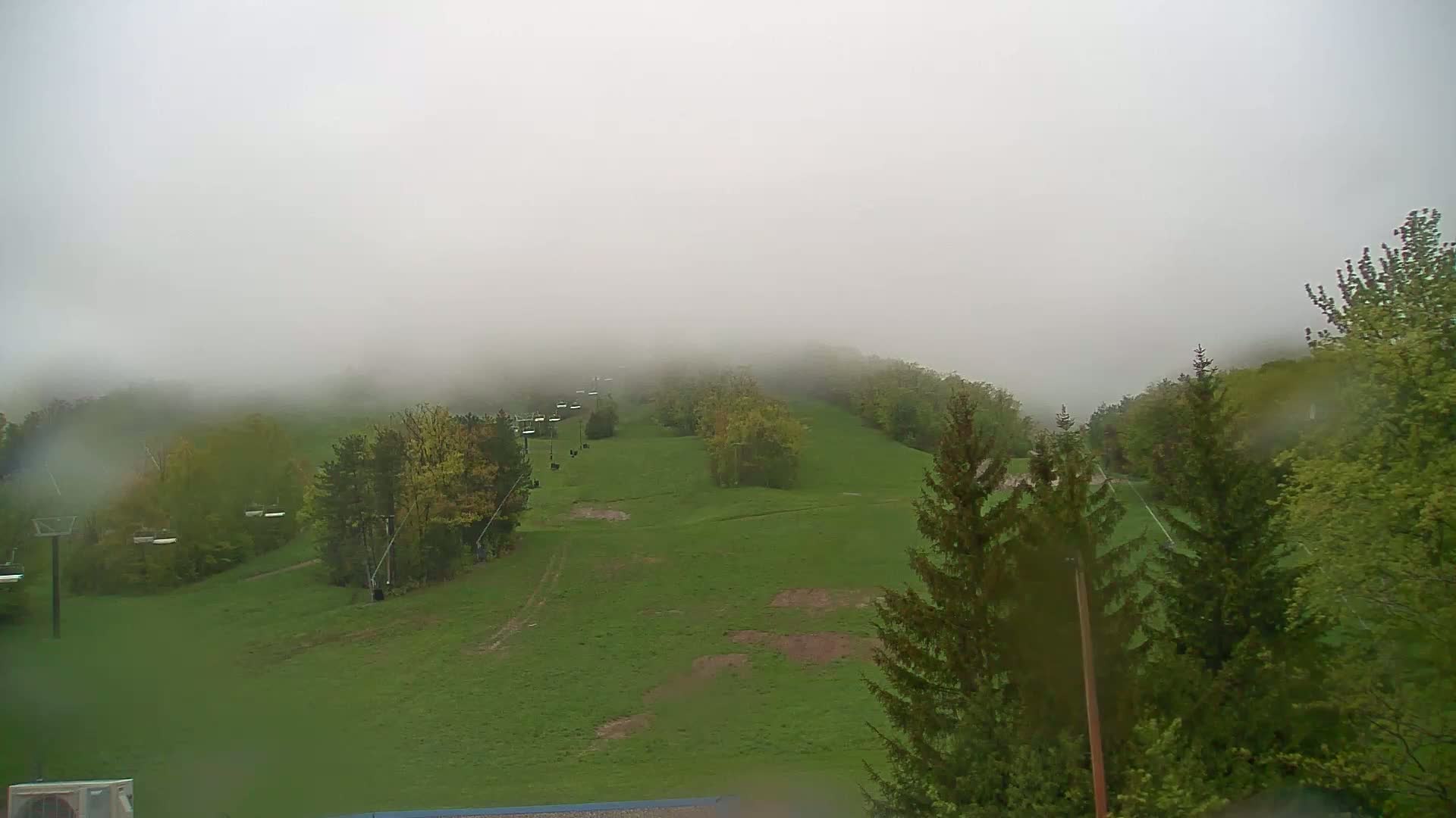 A foggy, rainy day reveals a grassy hillside with a ski lift and scattered trees.