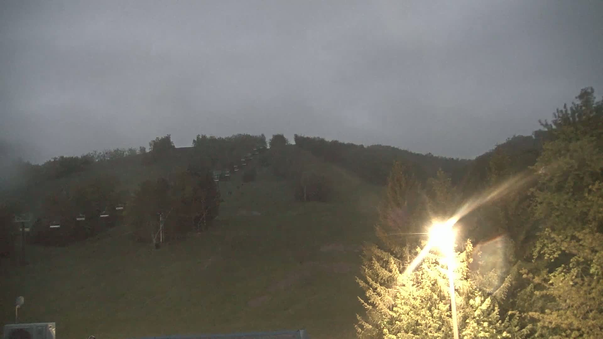 A dark, overcast sky hangs over a grassy hill dotted with trees and a ski lift.