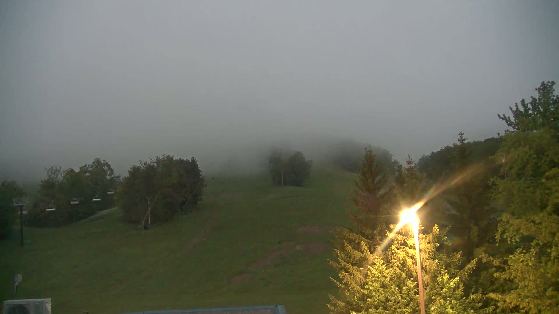 A foggy, grassy hillside with trees and a chairlift is illuminated by a streetlight.