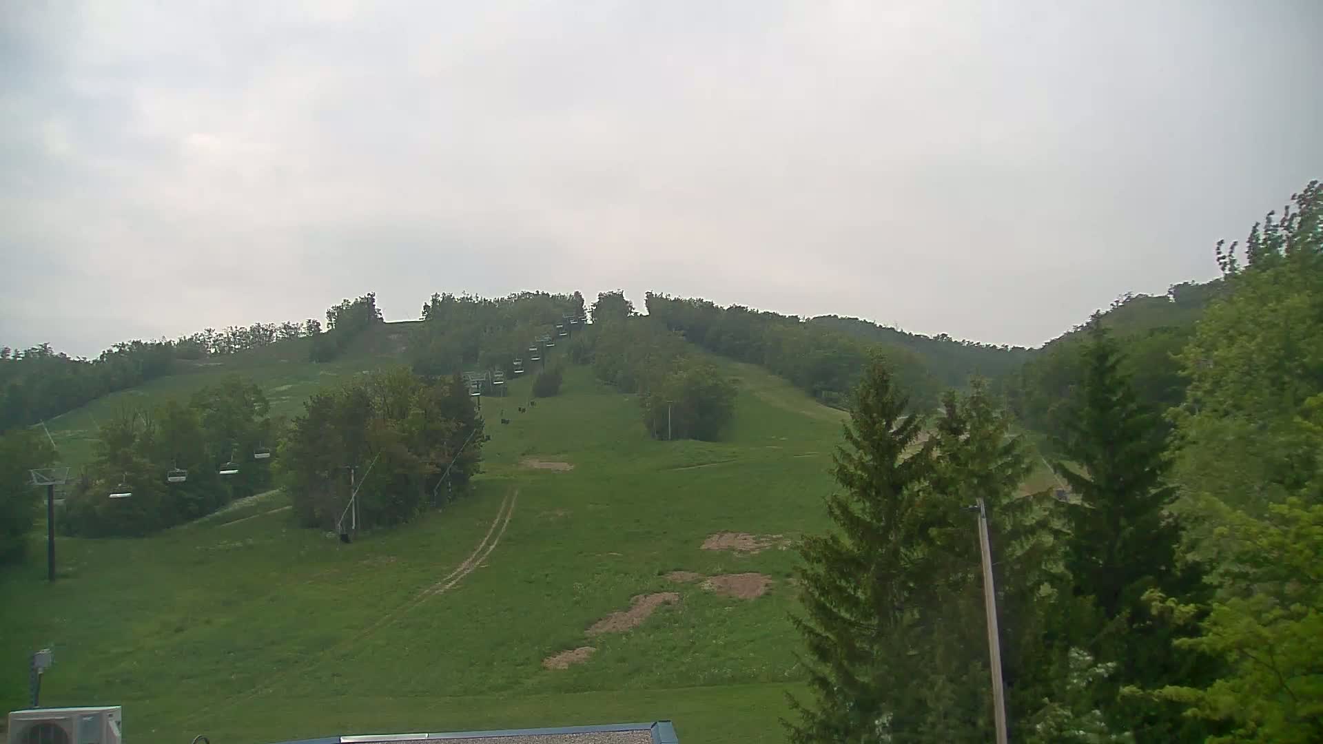 A grassy hillside with a chairlift and surrounding trees is visible under an overcast sky.