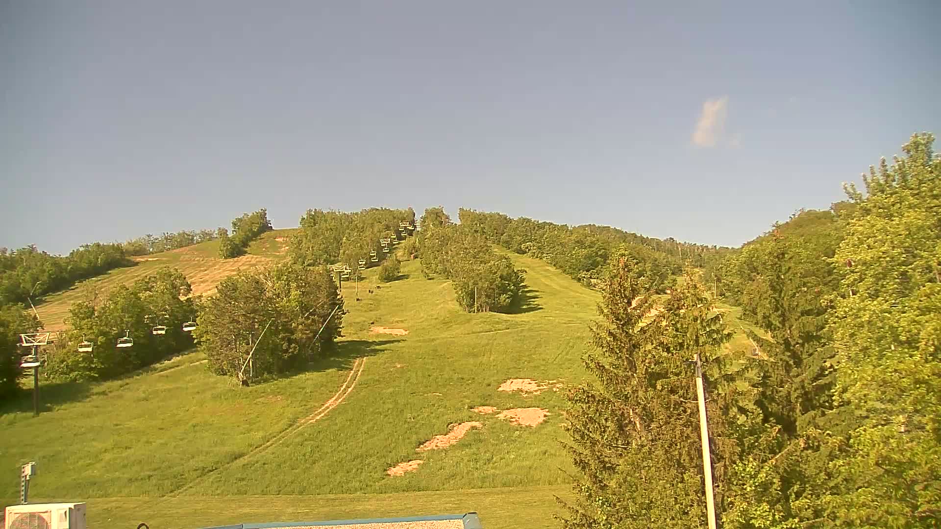 A grassy hillside with a chairlift ascending through a wooded area under a clear, sunny sky.