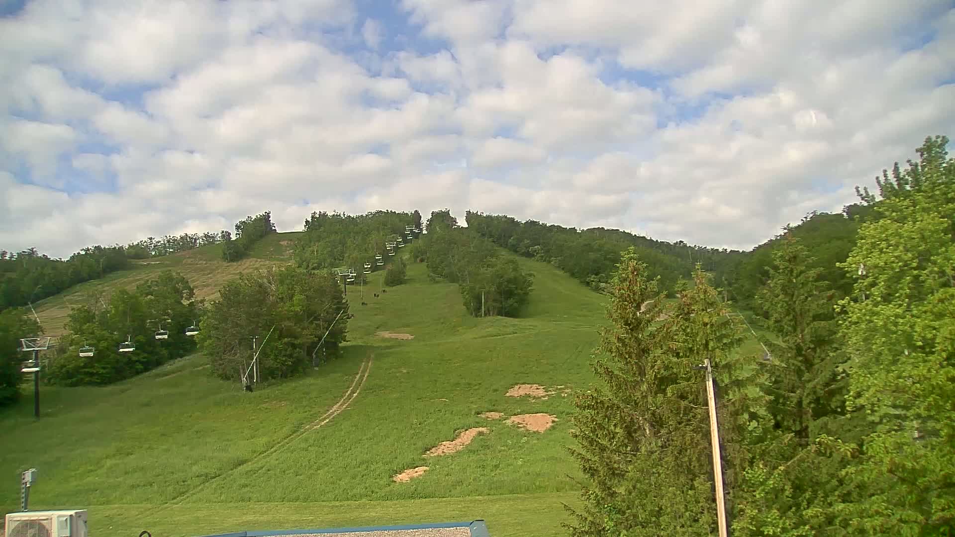 A grassy hillside with a chairlift ascending to a wooded peak under a partly cloudy sky.