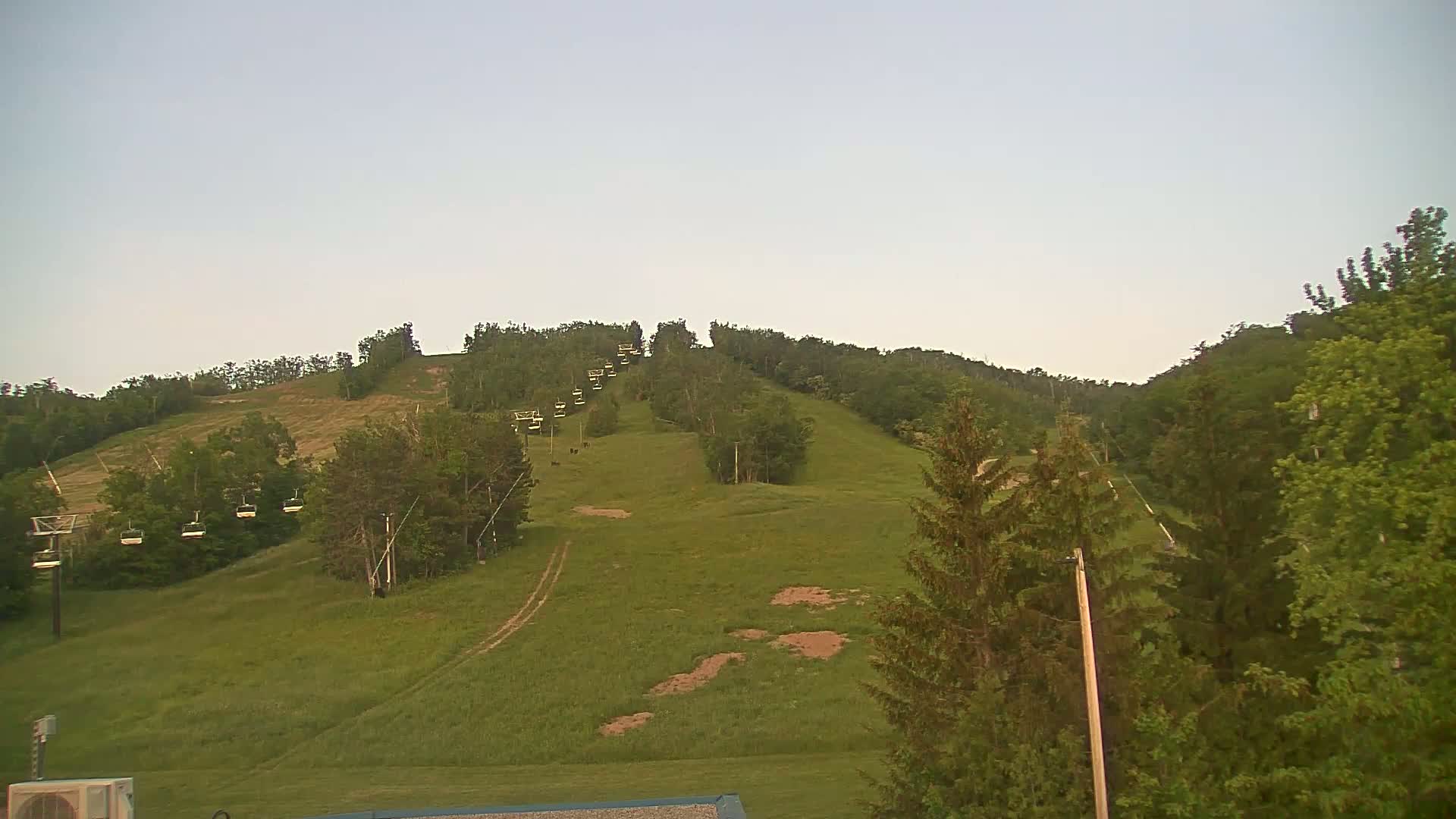 A chairlift ascends a grassy hill covered in green trees under a clear, light-grey sky.