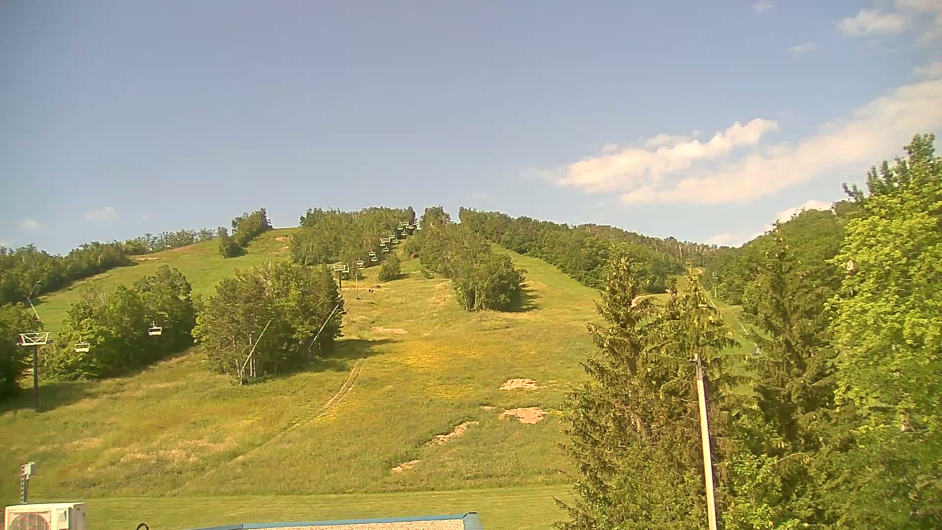 A grassy hill with a chairlift and scattered trees under a mostly sunny sky.