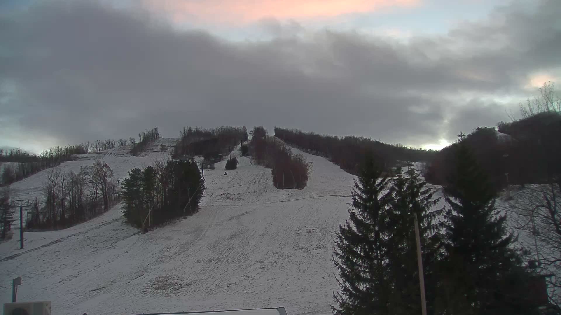 A snow-covered ski slope with scattered trees and visible ski lift poles is pictured under a heavily overcast, cloudy sky with a faint pink hue visible towards the upper left.