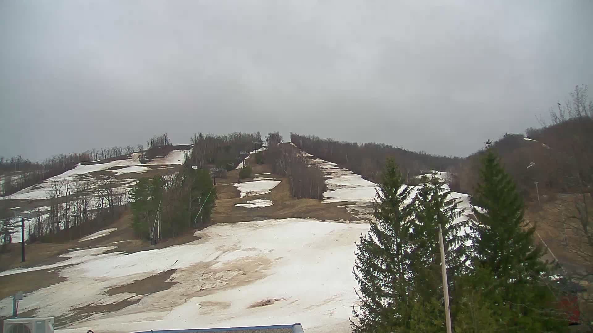 A snowy ski slope is visible at night, illuminated by a bright light source on the right amidst evergreen and bare trees, with what appears to be active snowfall.