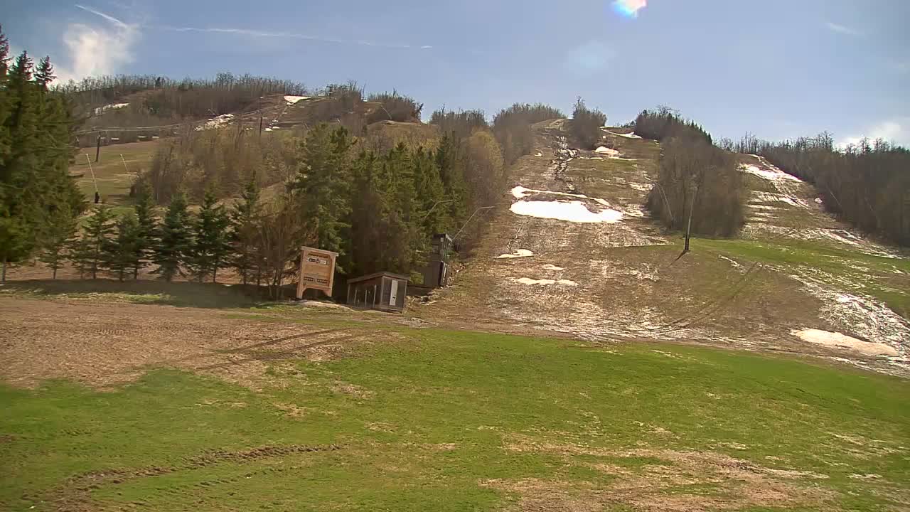 A mostly sunny day reveals a ski hill with melting snow patches, green grass emerging, and a small building near a row of evergreen trees.