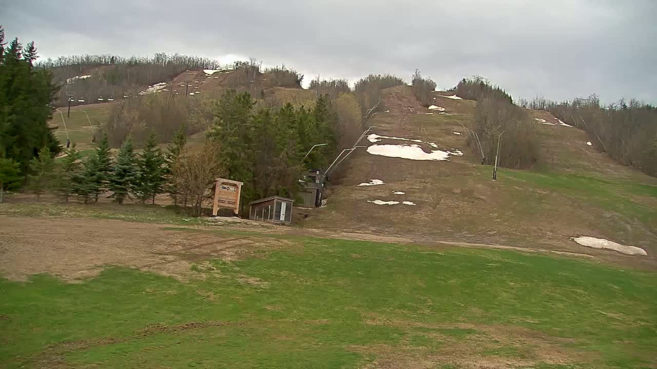 A mostly green grassy hill with patches of brown earth and small remnants of snow, featuring a ski lift and small structures, under an overcast sky.