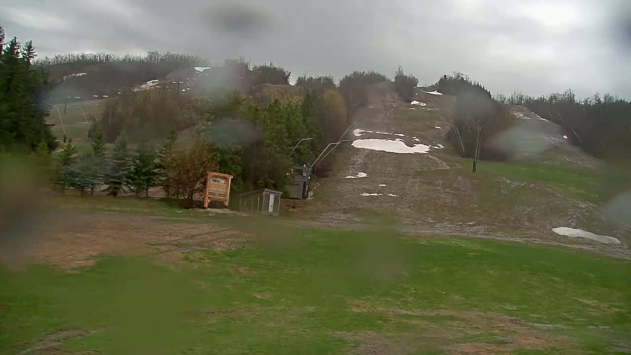 A rain-covered view of a ski hill in spring, showing patches of snow and green grass with a lift and small buildings at the bottom.