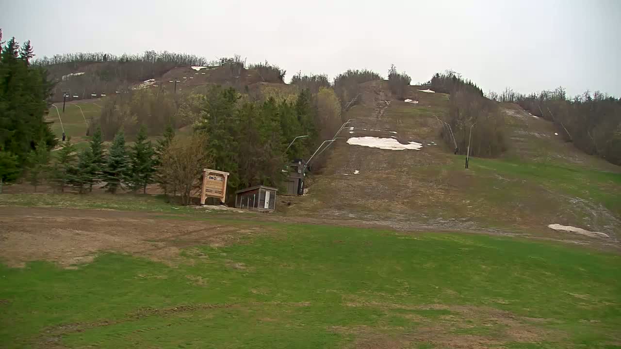 A mostly barren ski hill with patches of snow and new green grass shows a chairlift and small buildings under an overcast sky.