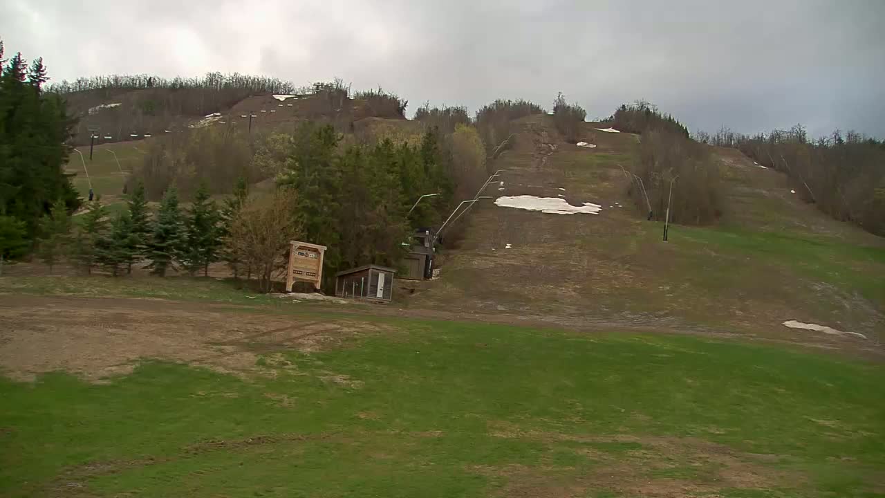 A mostly bare ski hill with patches of snow and new green grass under an overcast sky.