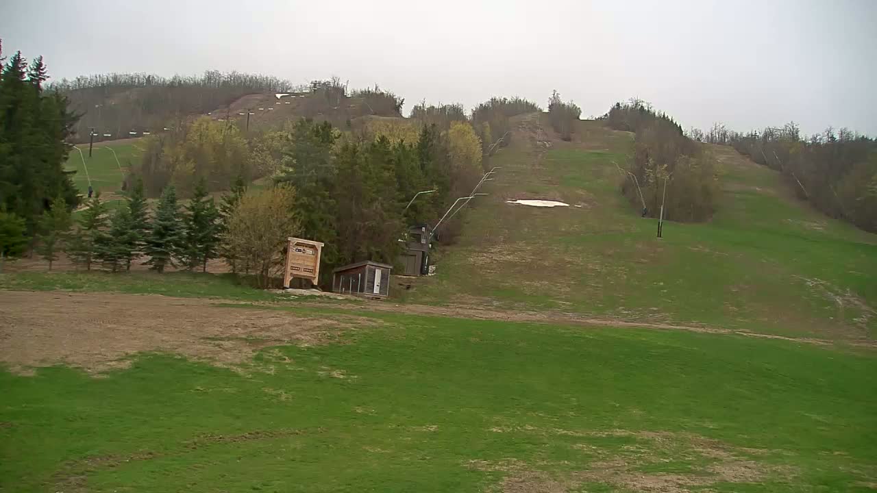 A mostly green grassy hillside with a few small buildings, a ski lift, and some remaining patches of snow under an overcast sky.