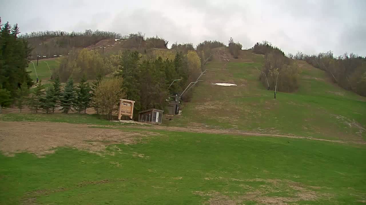 A grassy ski hill with a chairlift, small buildings, and a few patches of remaining snow under an overcast sky.