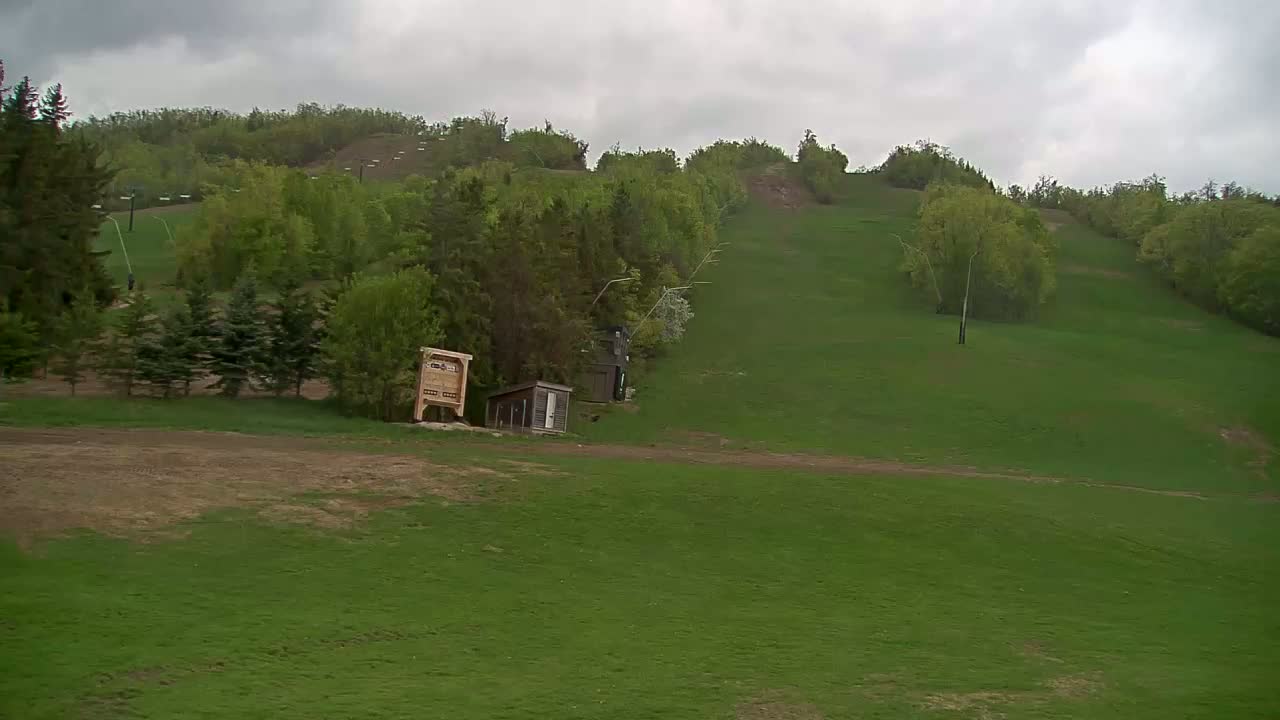 A grassy hill, under a cloudy sky, features a small building, a sign, and a ski lift in the background.