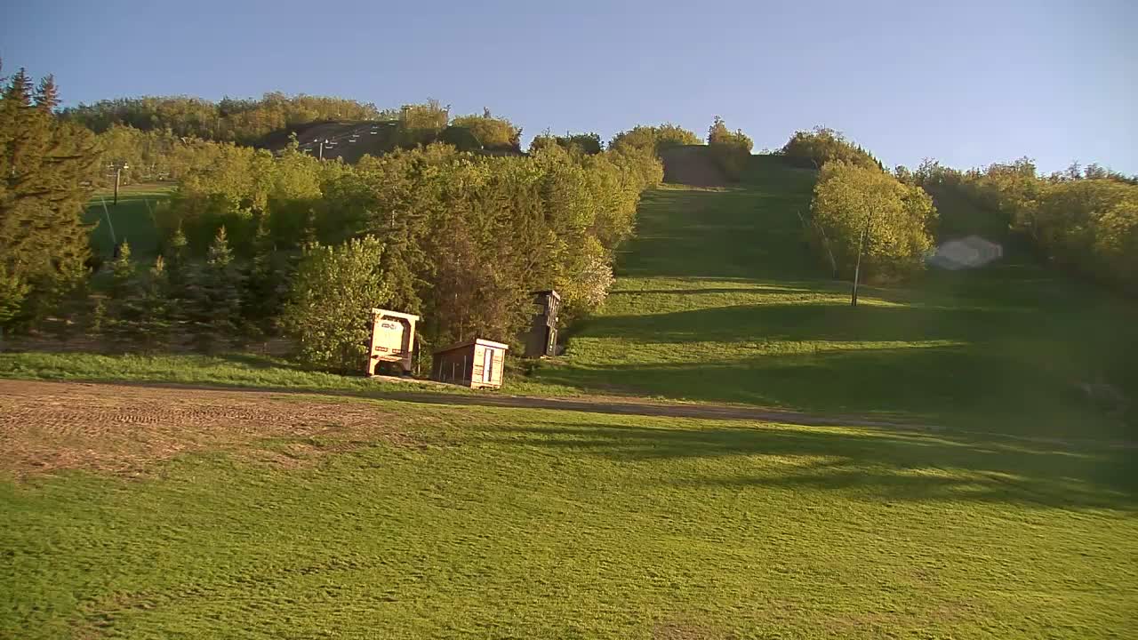 A grassy hill, dotted with small structures, slopes upward towards a tree-lined hill under a clear, sunny sky.