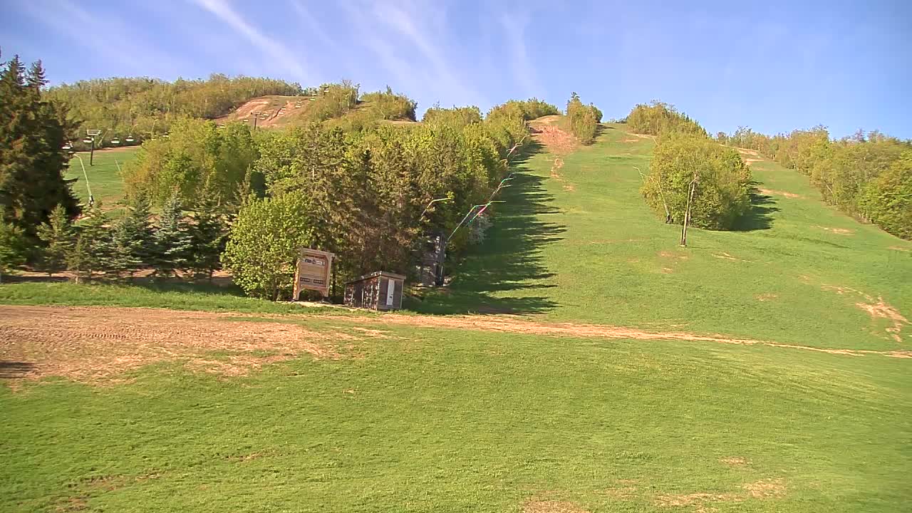 A grassy hill with a ski lift and small building under a mostly sunny sky.