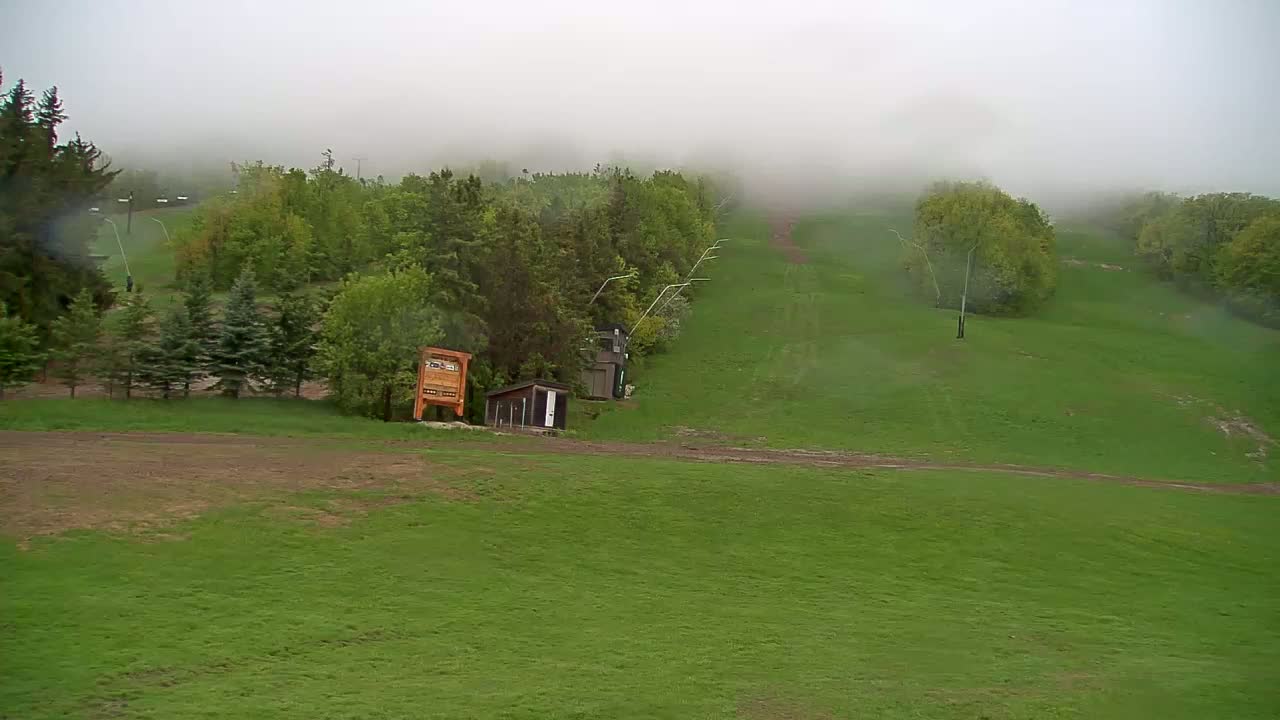 A grassy hill with a ski lift and small buildings is shrouded in fog and light rain.