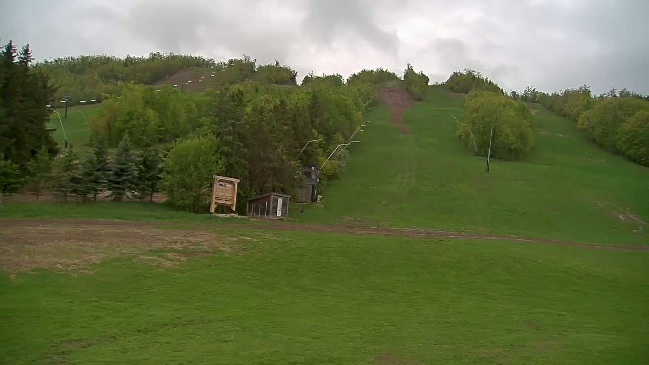 A grassy ski hill with a lift and small structures is visible under an overcast sky.