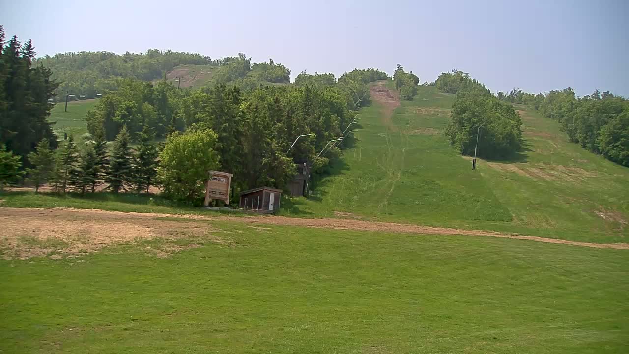 A grassy ski hill in the summer, with a small building and a lift visible in the mid-ground, under a clear sky.