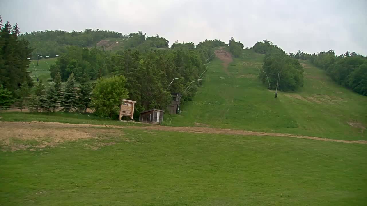 A grassy ski hill in overcast weather shows a chairlift and small buildings at the base of the hill.