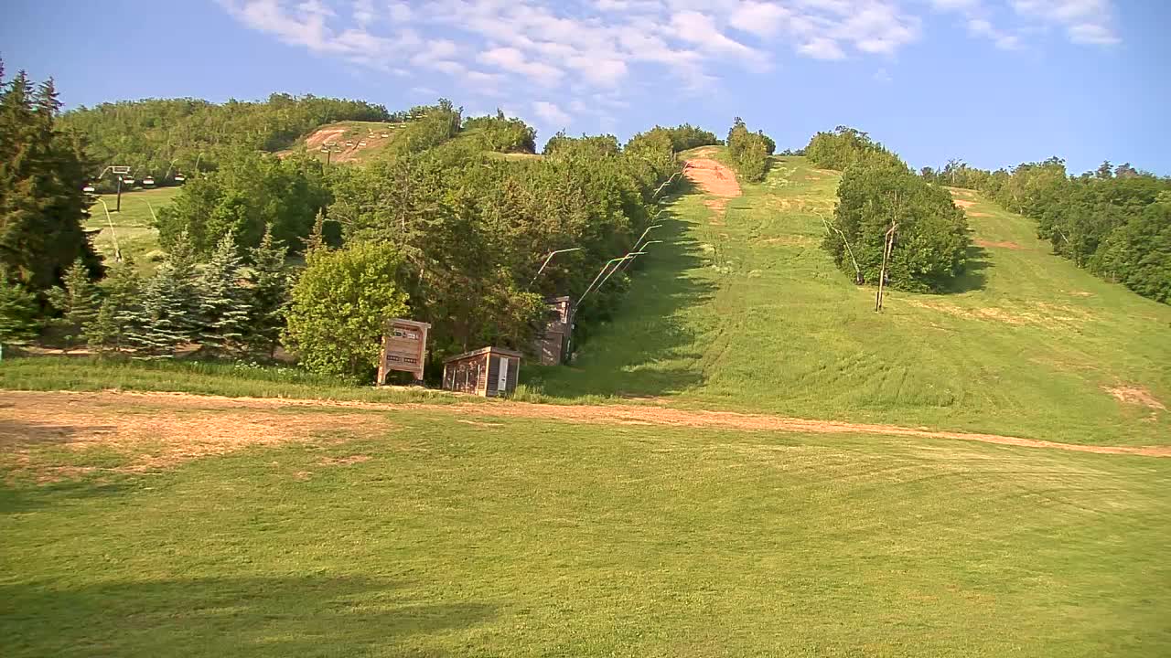 A grassy hillside with a ski lift and small buildings, under a partly cloudy sky.